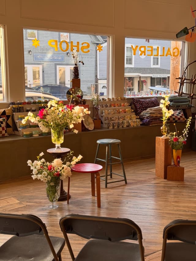 A sunlit shop interior with flower arrangements, merchandise displays, and chairs facing windows labeled "SHOP" and "GALLERY."