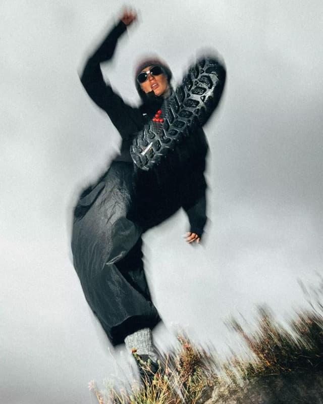 Motion-blurred person in dark clothing and sunglasses raises a leg, showcasing the sole of a hiking boot.