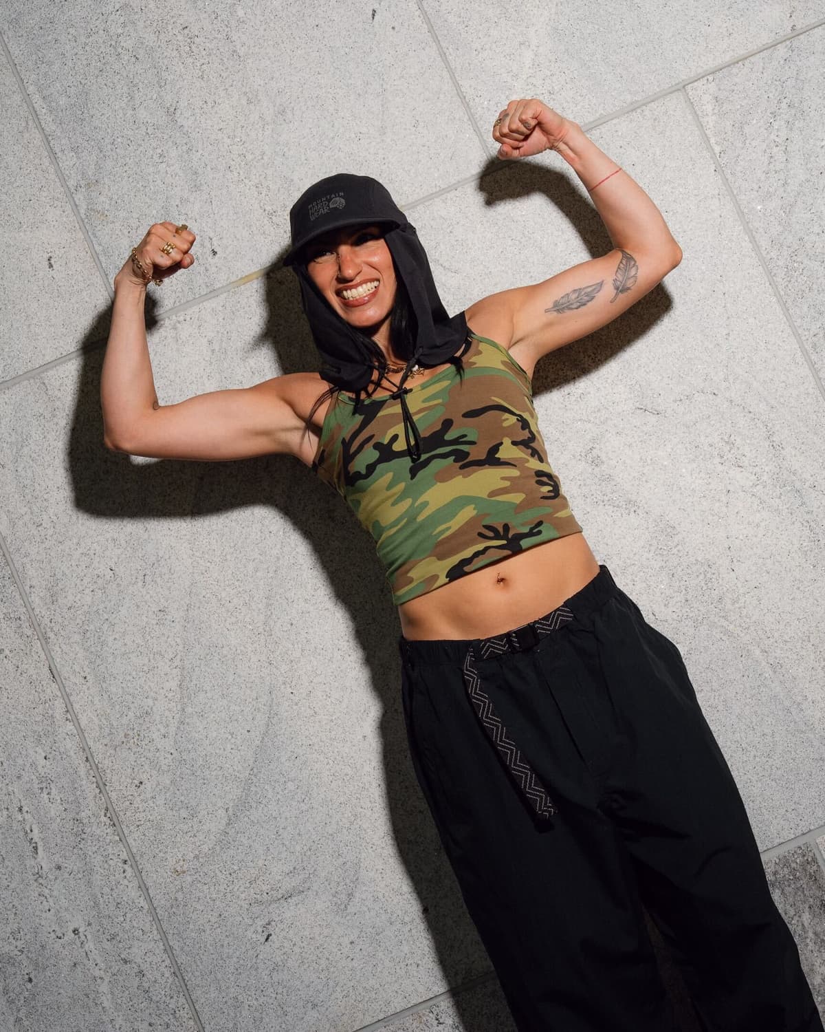 Smiling person in a camouflage crop top and black hat, flexing biceps against a gray wall.