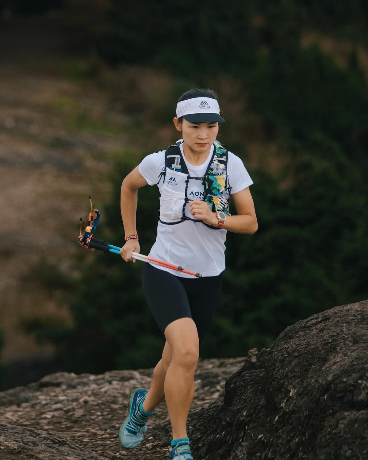 A female trail runner with a running vest and trekking poles runs on a rocky path.