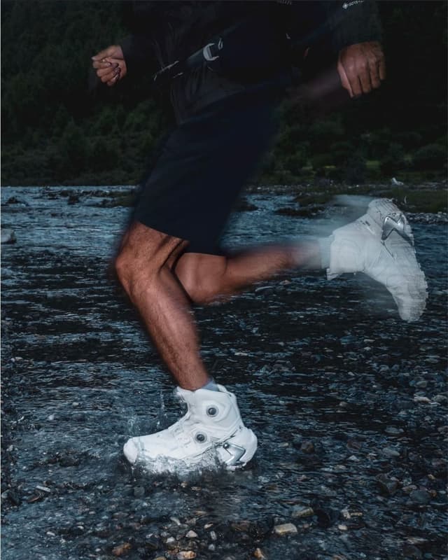 A person in black shorts and white shoes runs through shallow water, creating a splash.