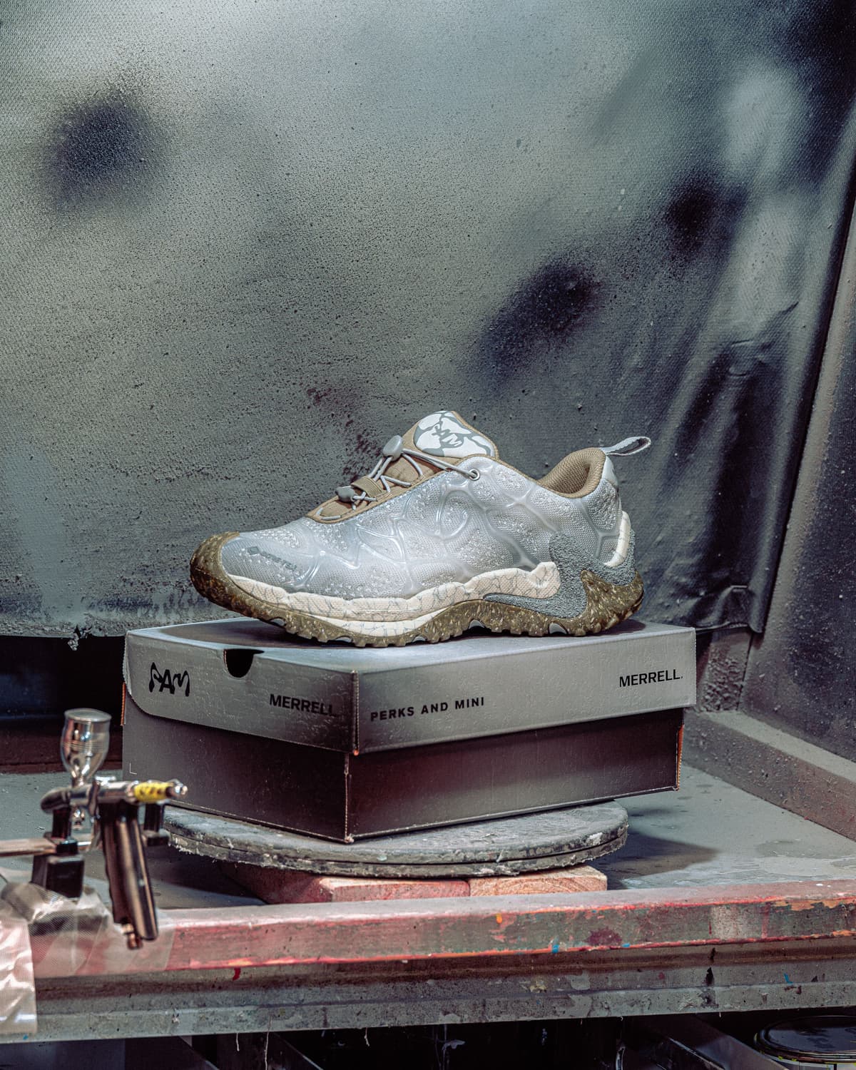 A white Merrell shoe with a translucent, patterned upper and speckled sole rests on a Merrell Perks and Mini box, an airbrush visible in a workshop setting.