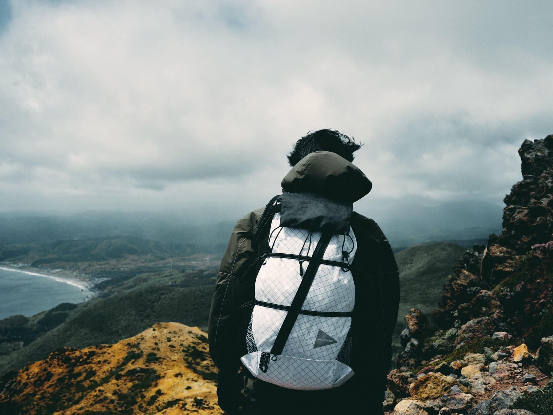 A person with a white backpack stands on a mountain peak, looking at a cloudy landscape with a coastline below.