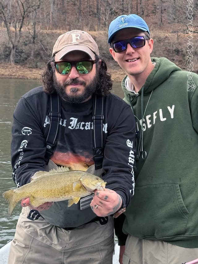 Two smiling men on a boat, one holding a smallmouth bass.