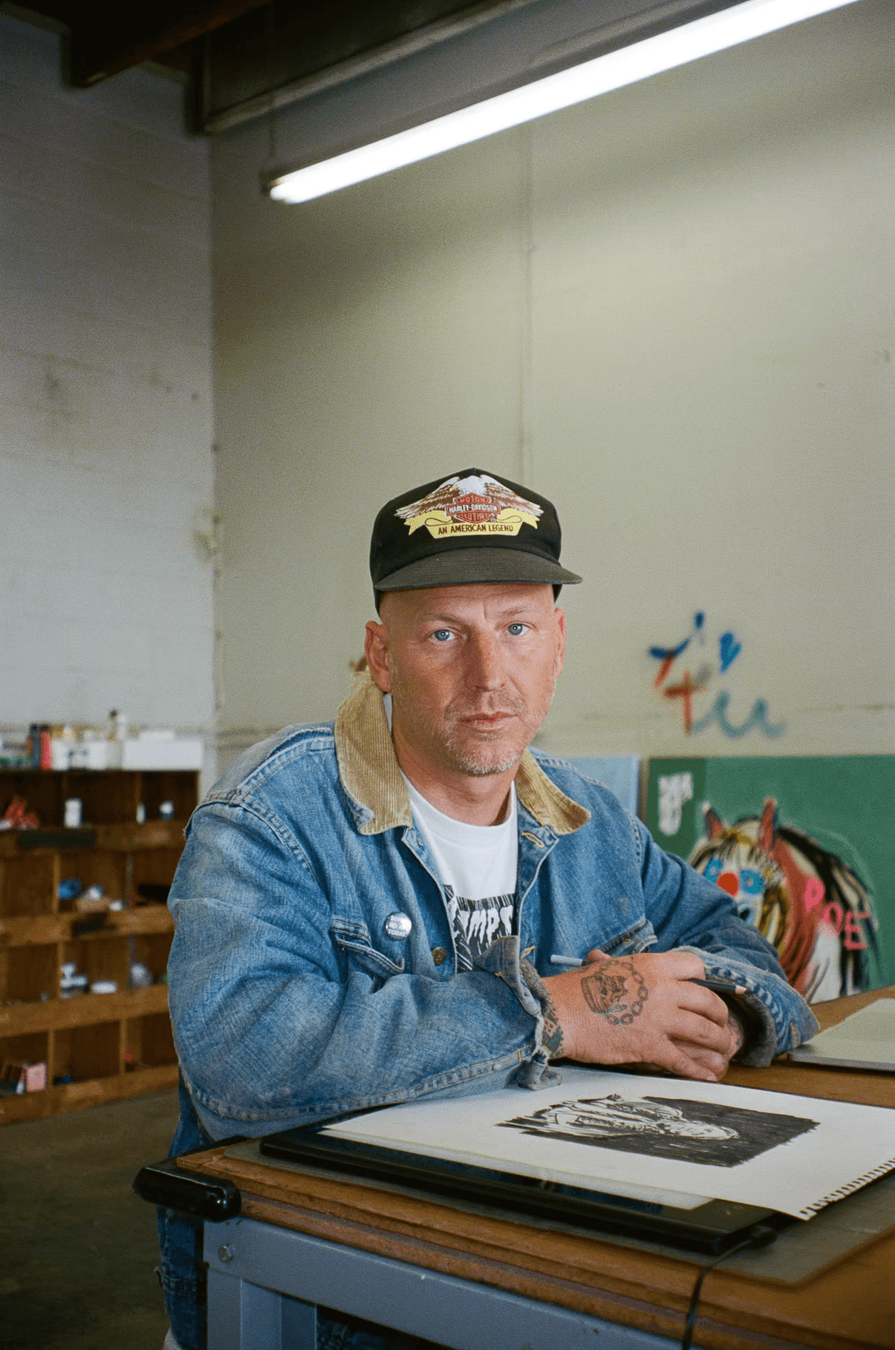 A man with a shaved head, Harley-Davidson cap, denim jacket, and tattooed hands sits at a desk in an art studio, looking directly at the viewer. A drawing is in front of him.