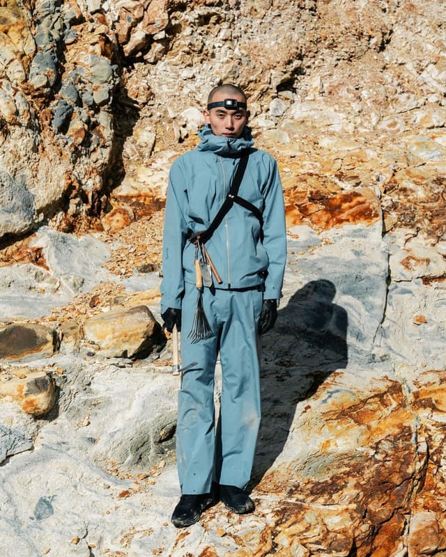 A man wearing a headlamp, blue outdoor suit, and tools stands against a rough, rocky cliff.