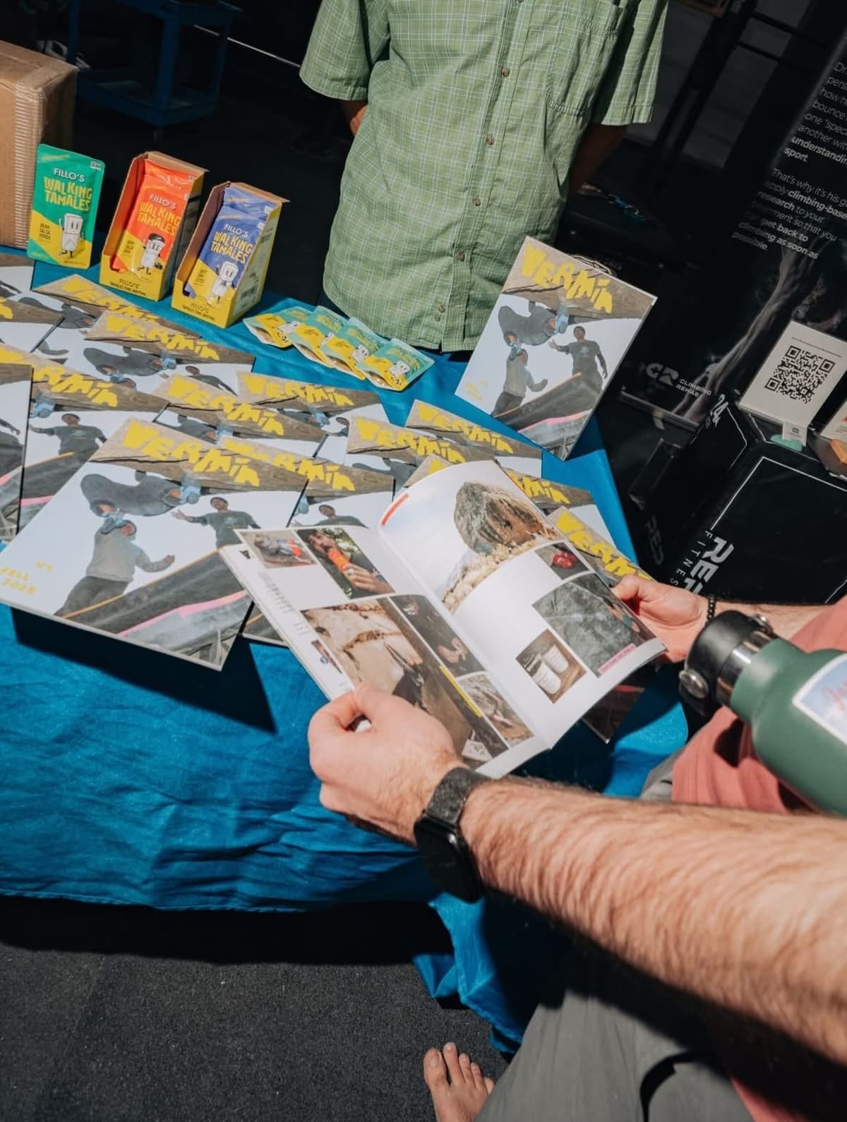 A person reads an open magazine at a blue table covered with numerous copies of a climbing-themed magazine, boxes of "Walking Tamales," and other items, with another person visible in the background.