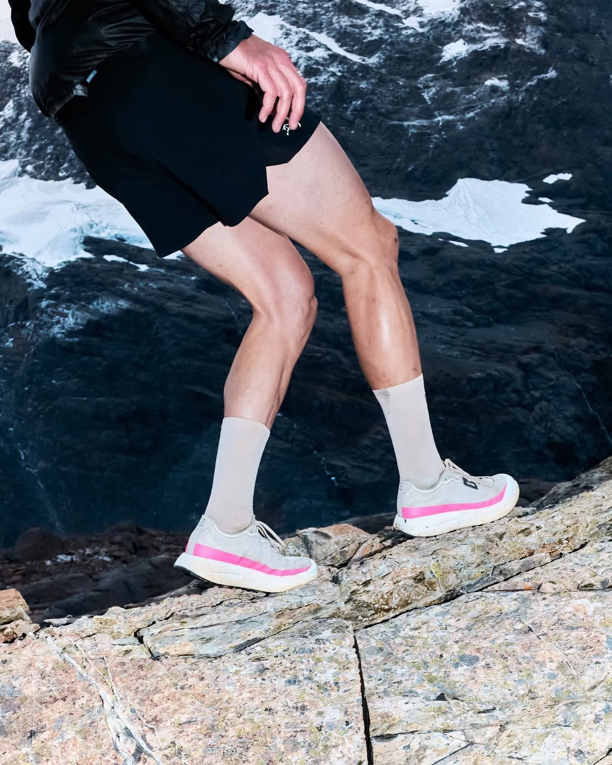 A person in black shorts and grey-and-pink trail shoes walks on a rocky surface with snowy mountains in the background.