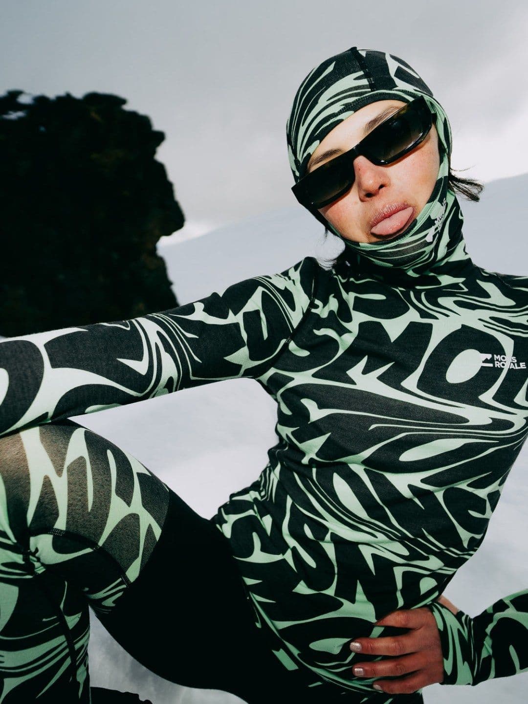 A person in a black and mint green Mons Royale patterned ski suit, balaclava, and sunglasses sticks their tongue out in a snowy landscape.