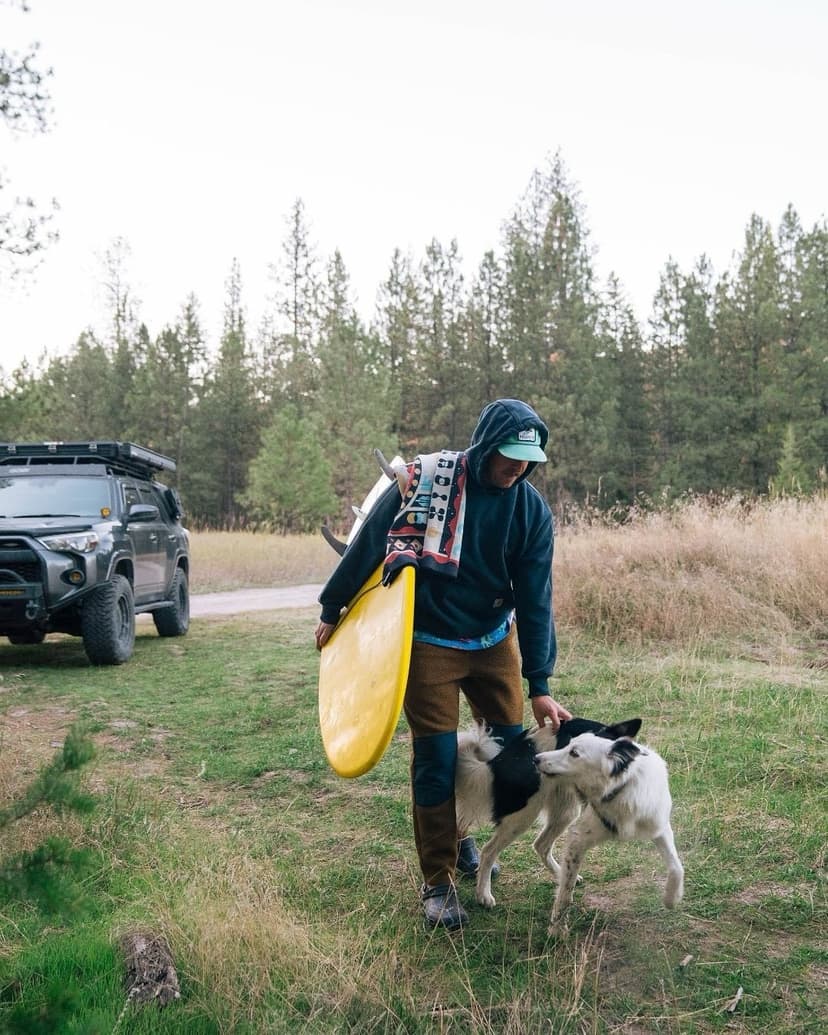 A person carrying a yellow boogie board and blanket pets a black and white dog in a grassy, wooded area with an SUV parked nearby.