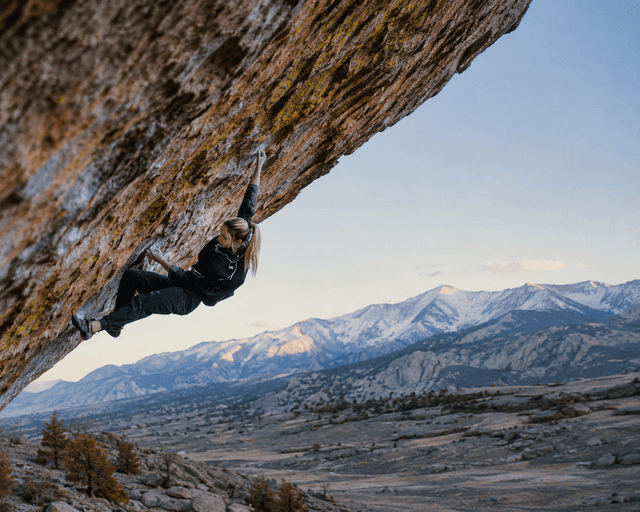 A woman bouldering on a rock overhang with snowy mountains and a valley in the background.