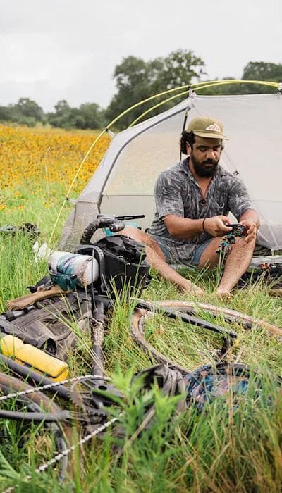 A bearded man in a hat sits barefoot by a small tent and bikepacking bicycle in a field of yellow wildflowers.