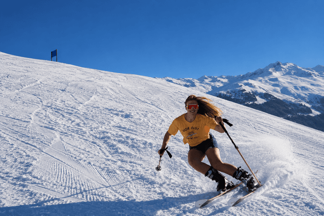A woman in a yellow t-shirt and shorts skis downhill, kicking up snow on a sunny mountain.