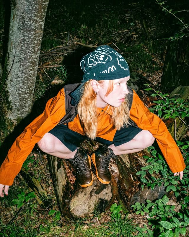 Person with long red hair, wearing a bandana, orange jacket, and hiking boots, crouching on a tree stump in a forest.