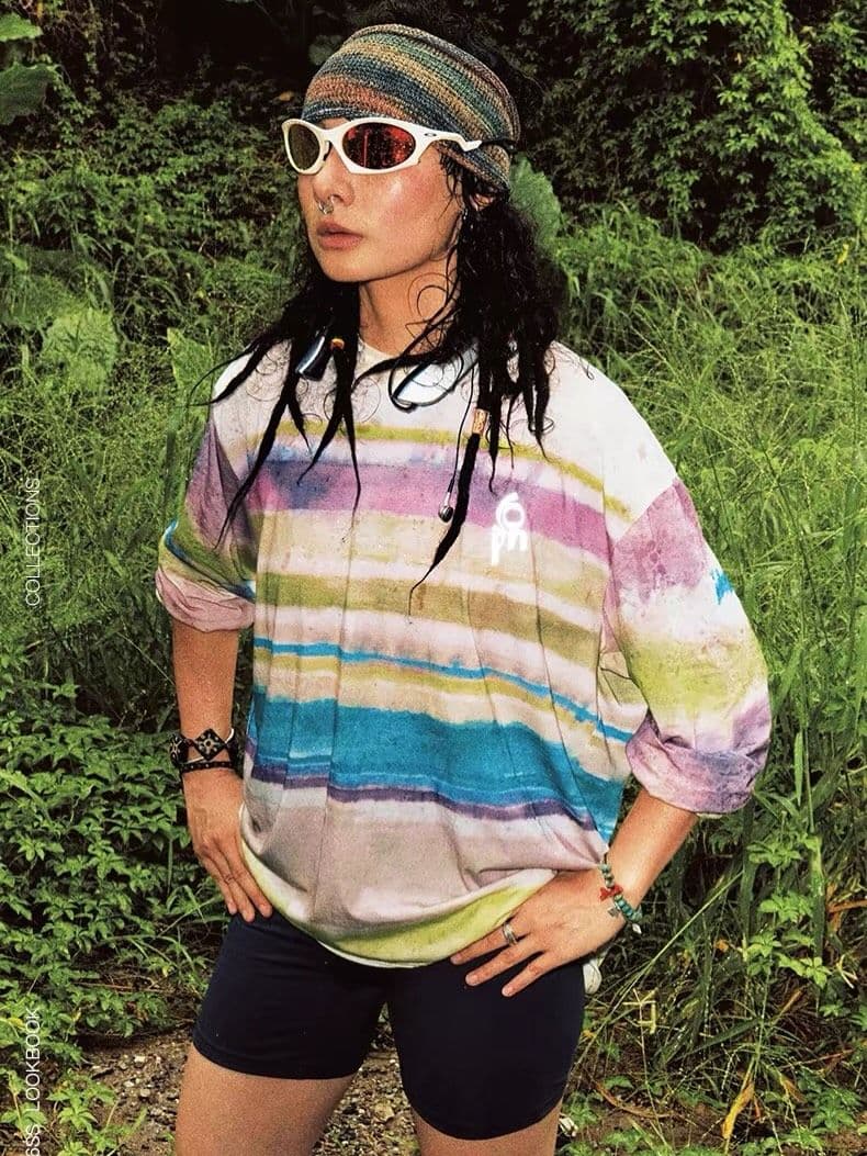 A person with a striped headband and white sunglasses wears an oversized pastel striped t-shirt and dark shorts, standing in lush green foliage.