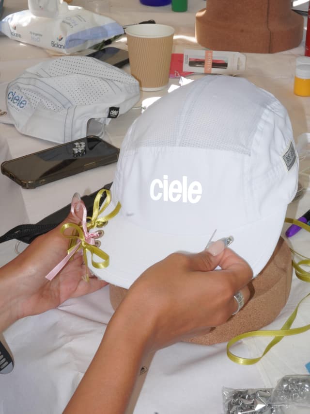 Hands decorate a white Ciele running cap with pink and green ribbons on a cork stand.