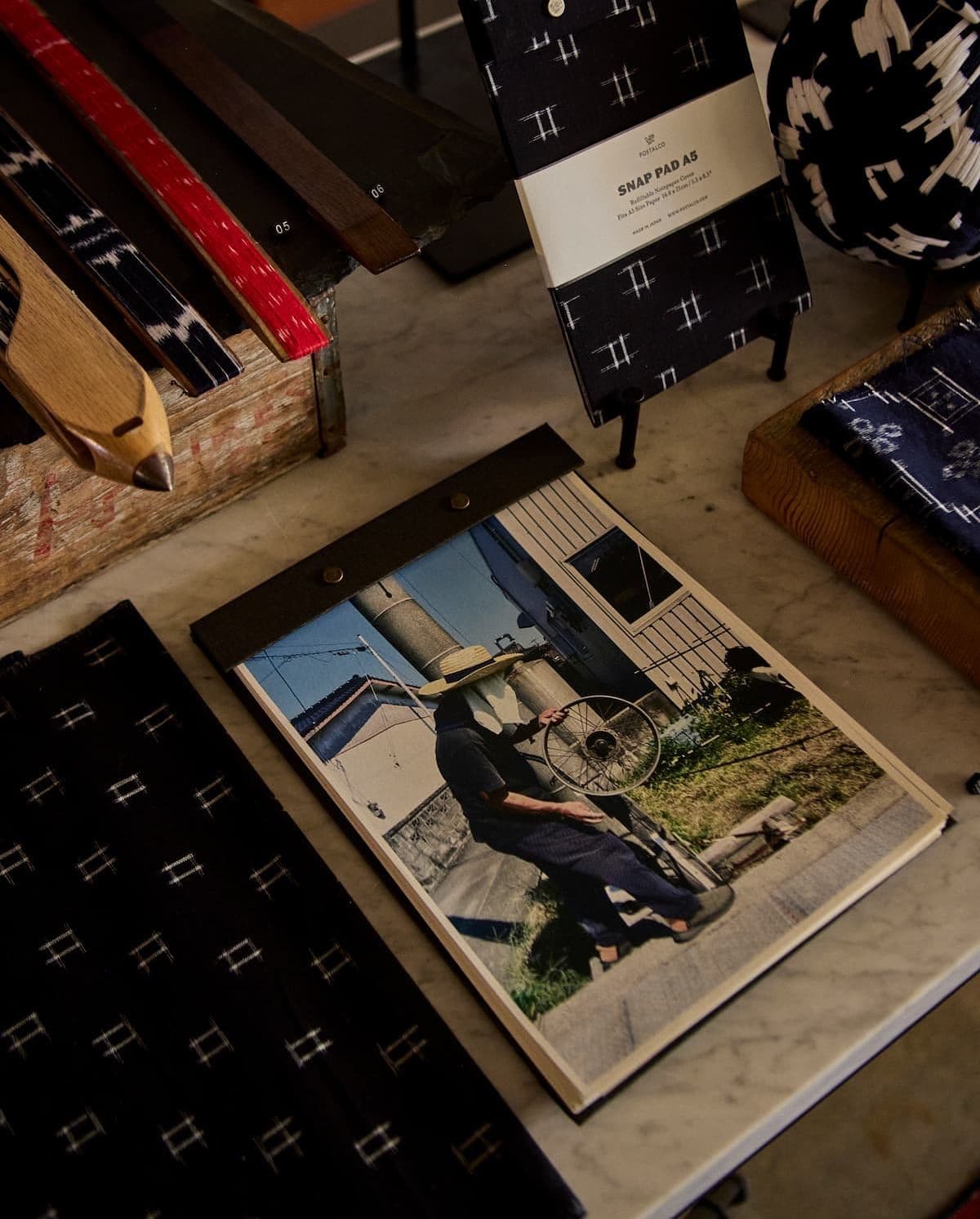 A marble table displays an open notebook with a photo of a person in a straw hat holding a bike wheel, alongside patterned textiles, a weaving shuttle, and a black patterned snap pad.