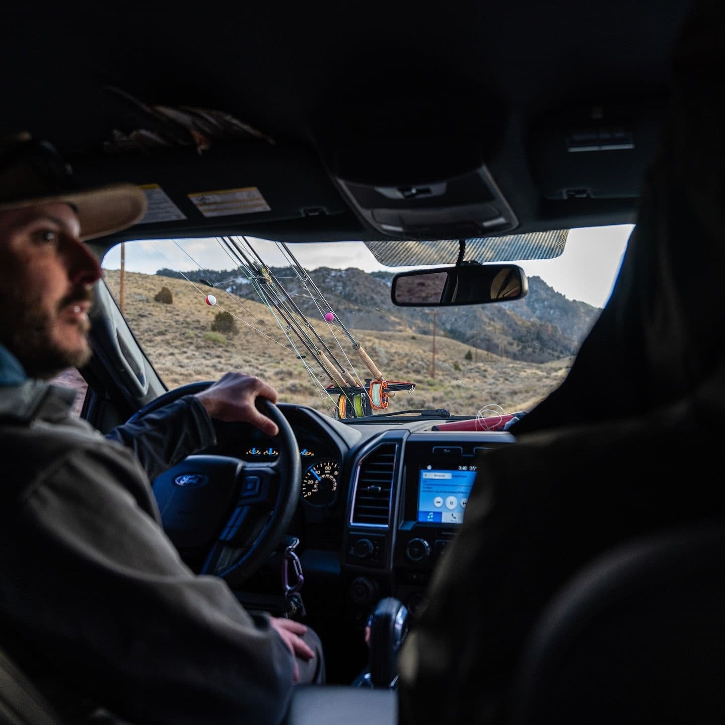 A man driving a truck with fishing rods mounted across the dashboard, looking out at a mountainous desert landscape.