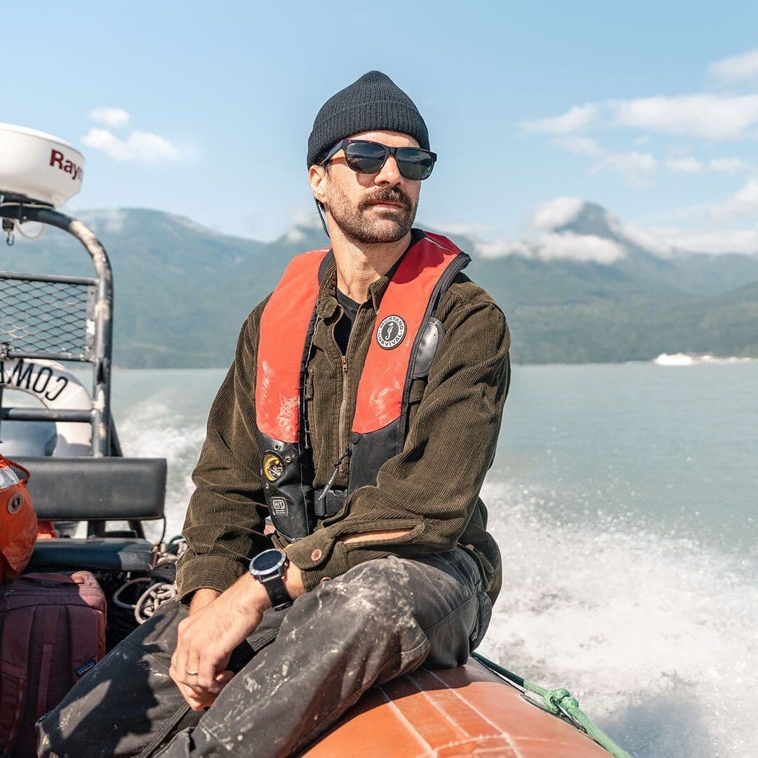 A man in a black beanie, sunglasses, and orange life vest rides in a boat on a sunny day with mountains and water in the background.