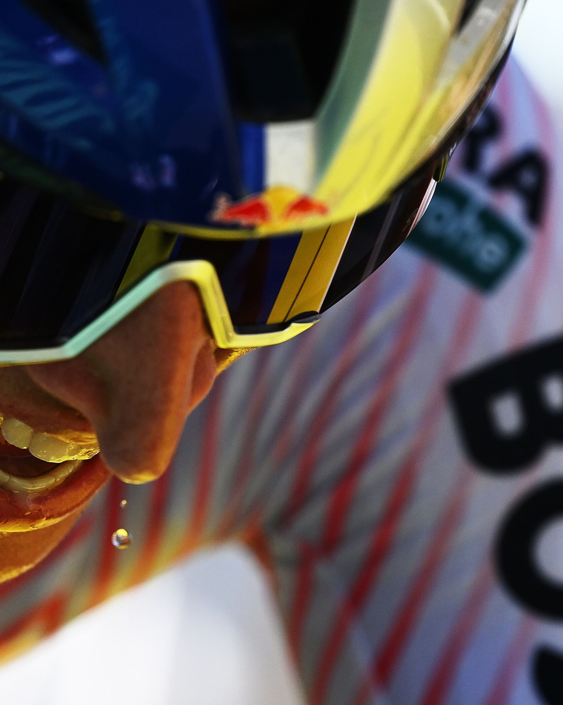A low-angle close-up of a cyclist wearing a blue helmet with a Red Bull logo and reflective sunglasses, with a drop of liquid falling from their chin.
