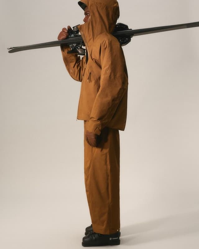 Person in brown hooded ski suit and black ski boots, carrying skis over their shoulder.