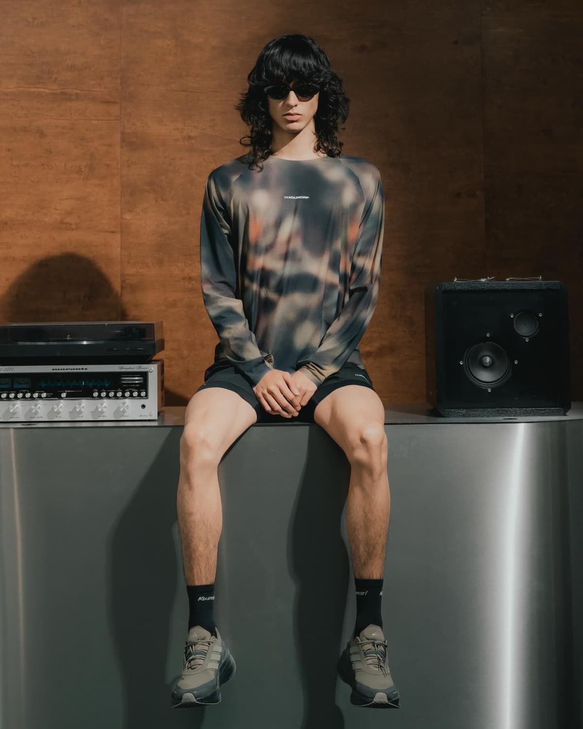 A person with long dark hair and sunglasses sits on a metal surface, wearing a patterned long-sleeve shirt, shorts, and sneakers, with vintage audio equipment nearby.