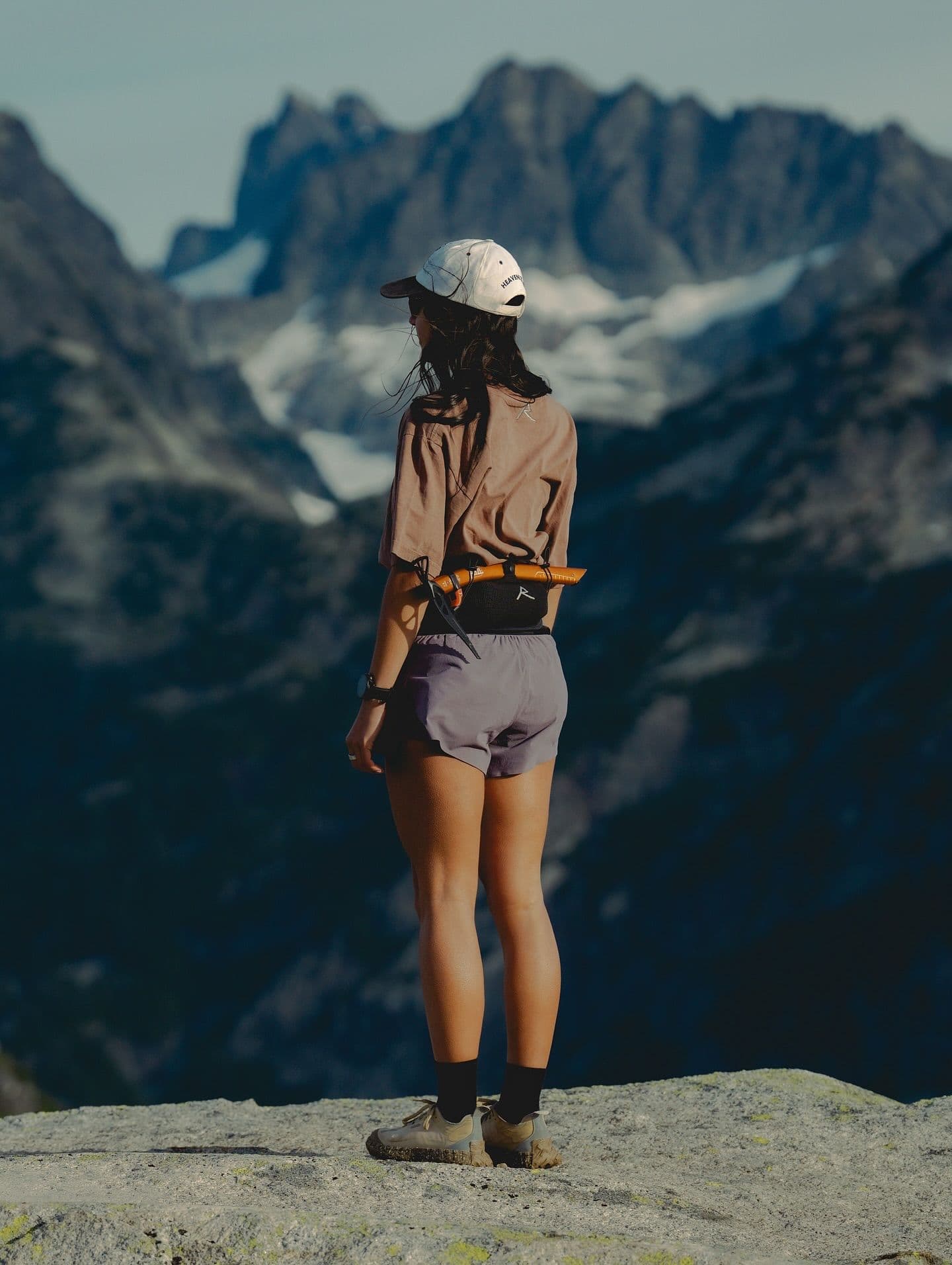 A person in a cap and shorts stands on a rocky outcrop, back to the viewer, gazing at vast snow-capped mountains.