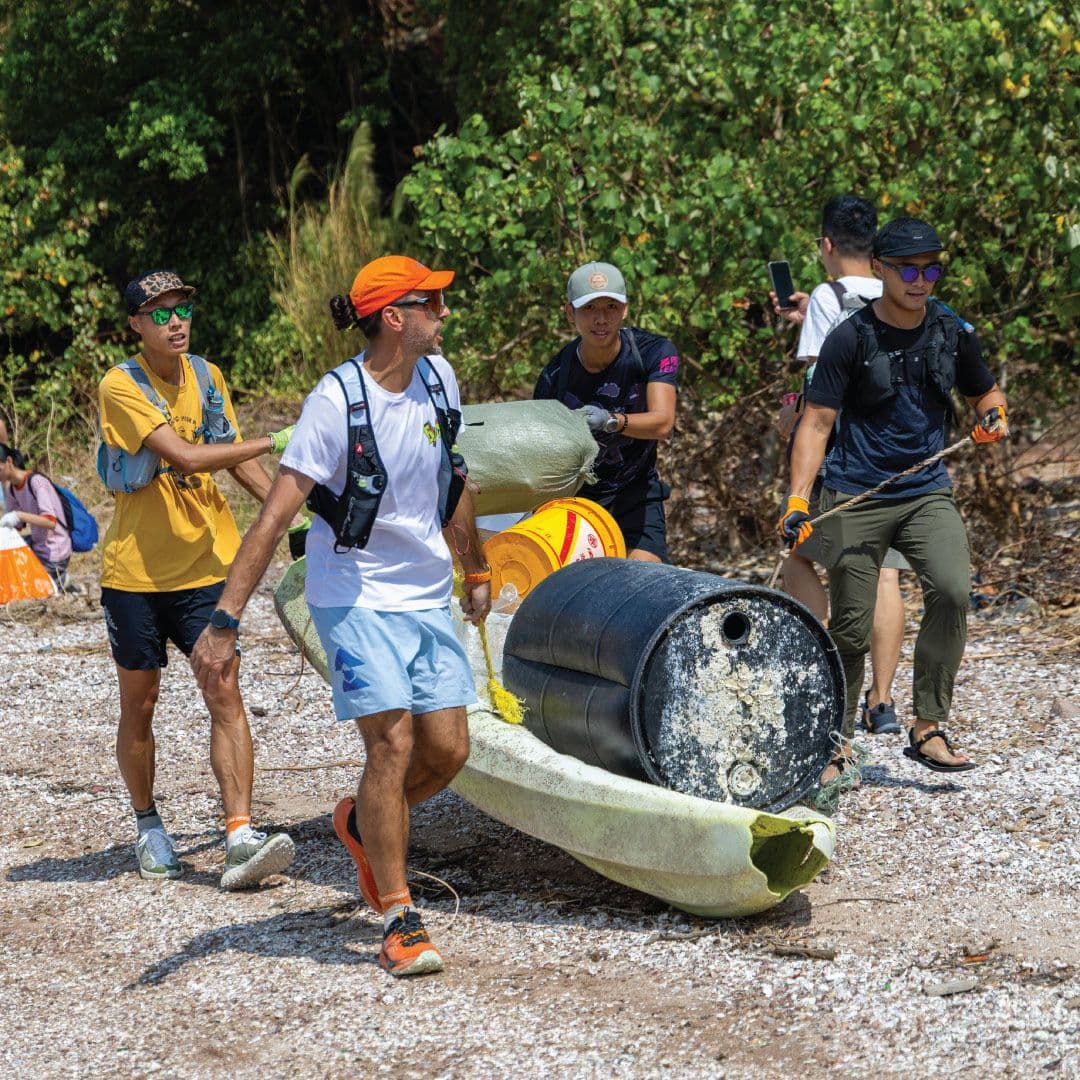 Volunteers carry large marine debris, including a broken kayak and barrels, from a rocky beach.