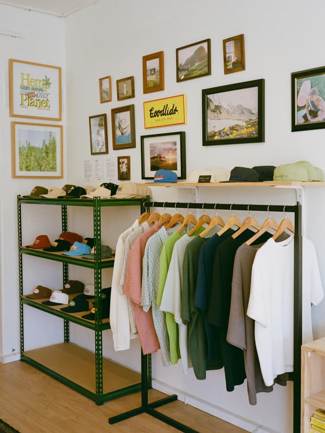 Interior of a bright clothing store featuring framed art on a white wall, a rack of colorful shirts, and a green shelving unit displaying hats.