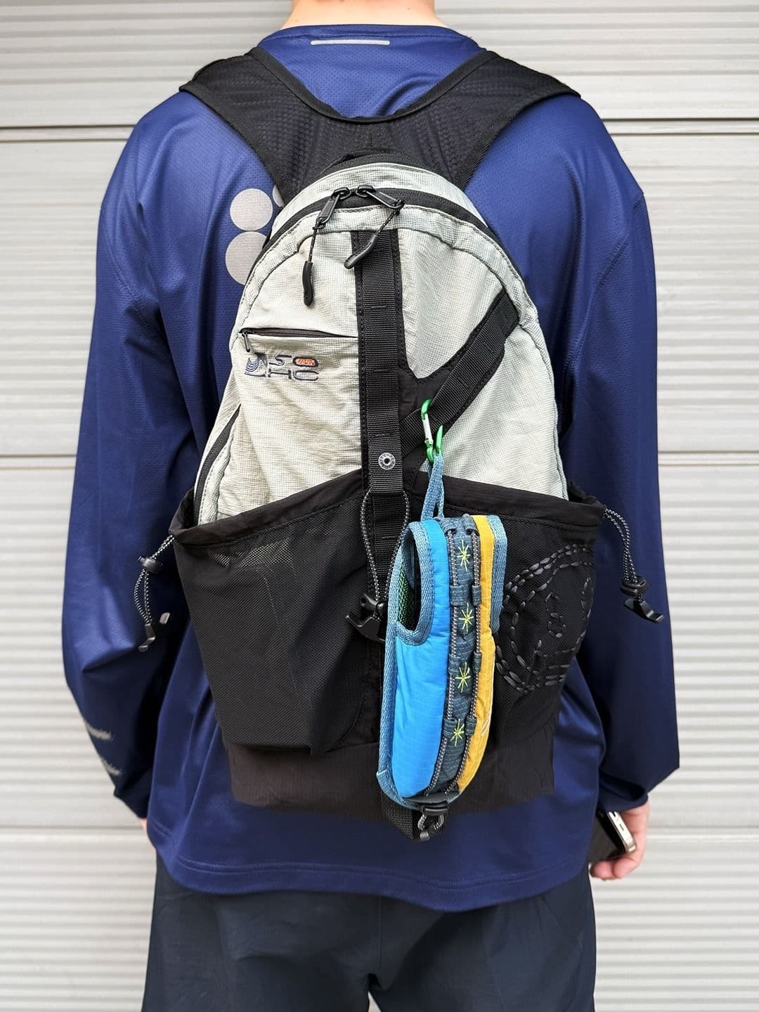 Back of a person wearing a navy shirt and a light grey backpack with a blue and yellow pouch attached.