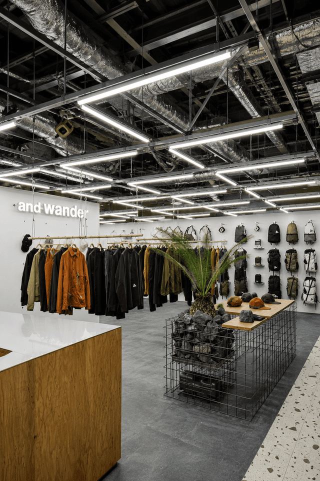 Modern clothing store interior with "and wander" logo, racks of apparel, backpacks on a wall, and exposed industrial ceiling.