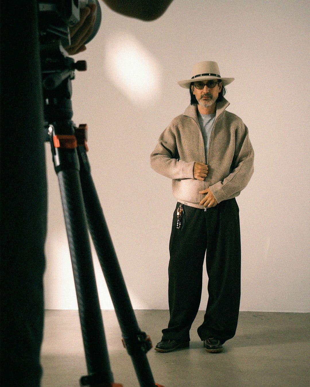 A man in a fedora, sunglasses, and beige zip-up jacket poses in a studio while being photographed.