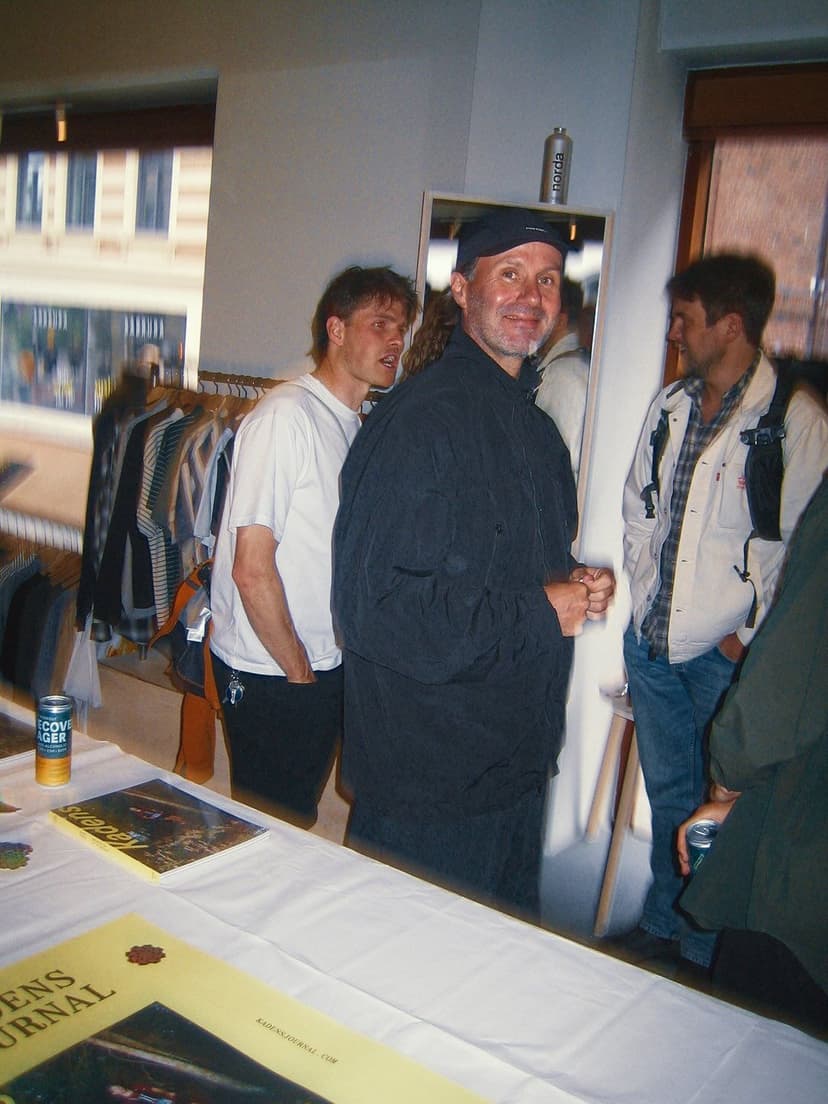Four men gathered indoors around a table with magazines and drinks, with one man in a black jacket and cap smiling at the camera.
