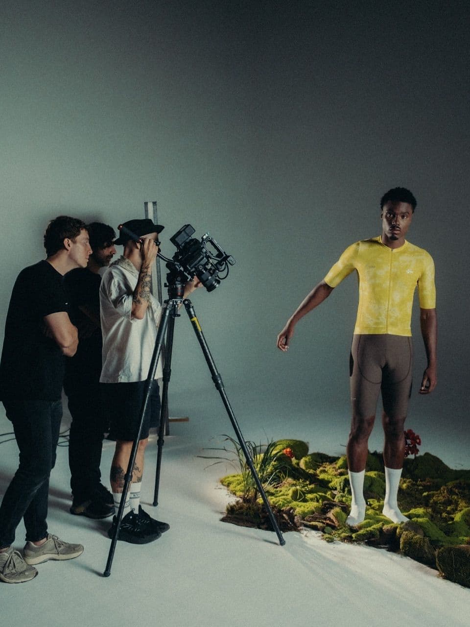 A film crew captures a male model in a yellow cycling jersey and brown shorts standing on a mossy set in a studio.