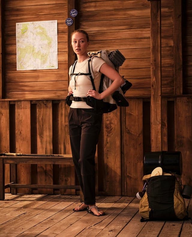 A woman wearing a large hiking backpack stands in a rustic wooden cabin with a map and trail signs on the wall.