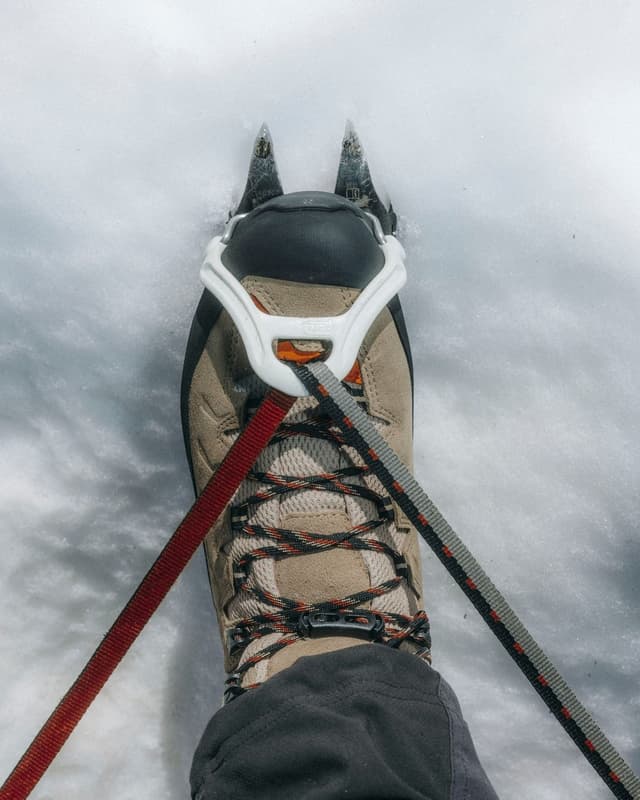 A hiking boot with front crampons and straps stands in snow.