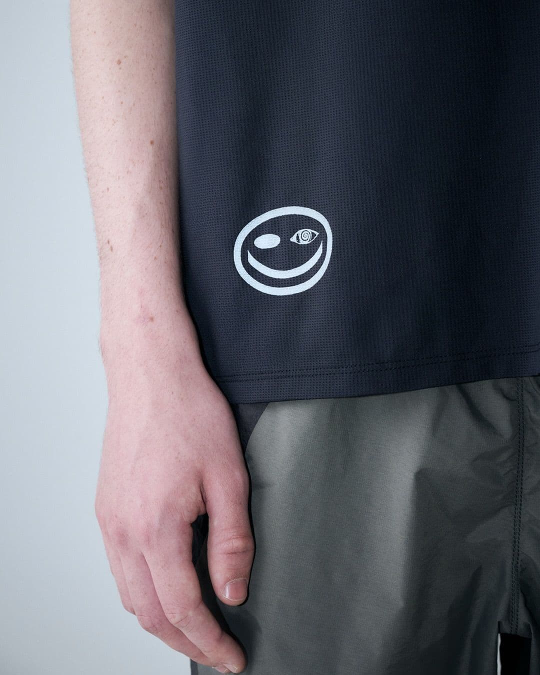 Dark grey shirt with a white logo of a winking smiley face with a spiraled eye, worn over dark grey pants.