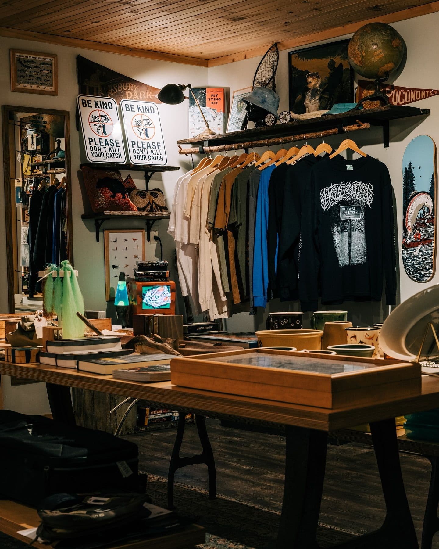 A rustic retail space with a wooden table displaying books and craft items in the foreground, and a clothing rack beneath shelves adorned with fishing gear, a globe, and "Be Kind" signs in the background.