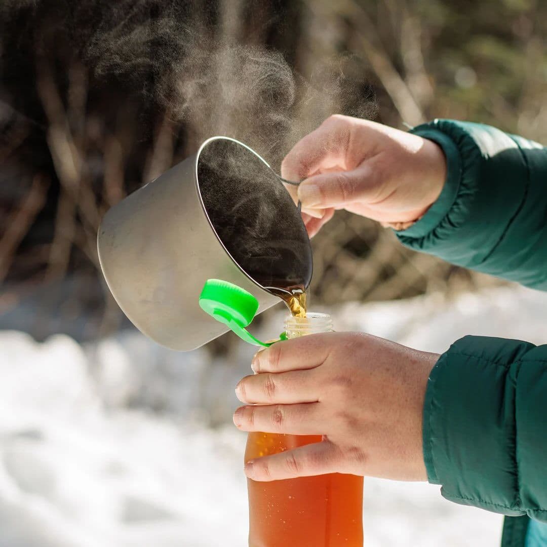 Hands pour steaming dark liquid from a pot into a bottle with a green cap in the snow.
