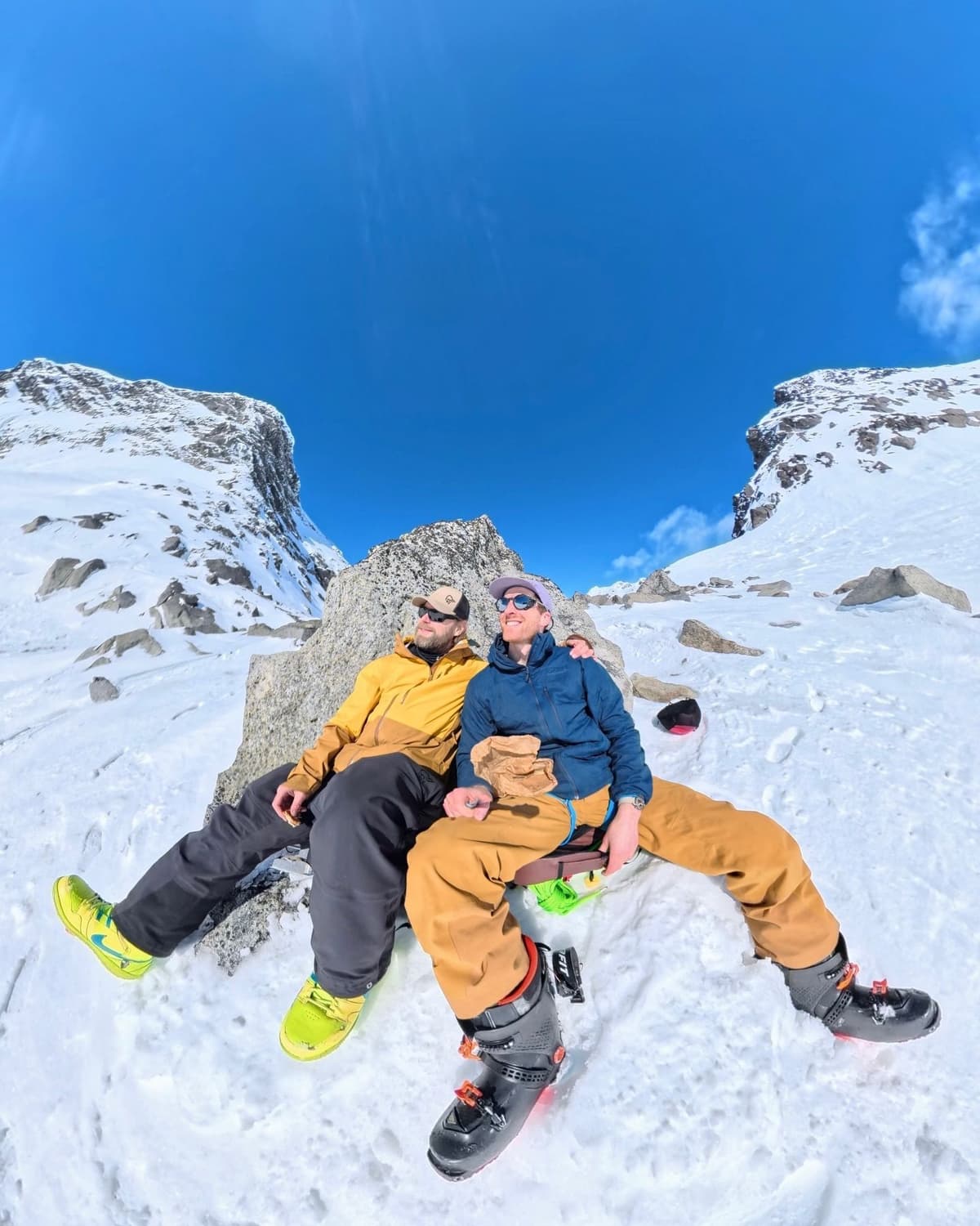 Two men, dressed for winter, sit on a rock in a snowy mountain landscape under a clear blue sky.