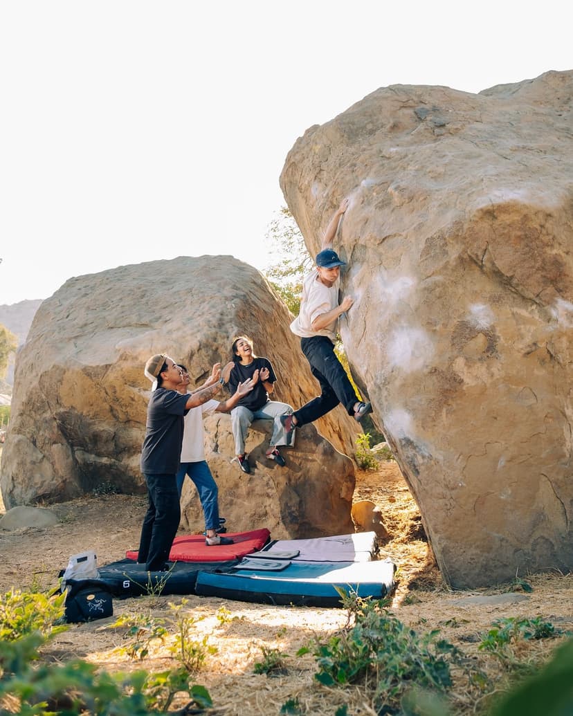 A person bouldering on a large rock, with three others watching and crash pads on the ground.