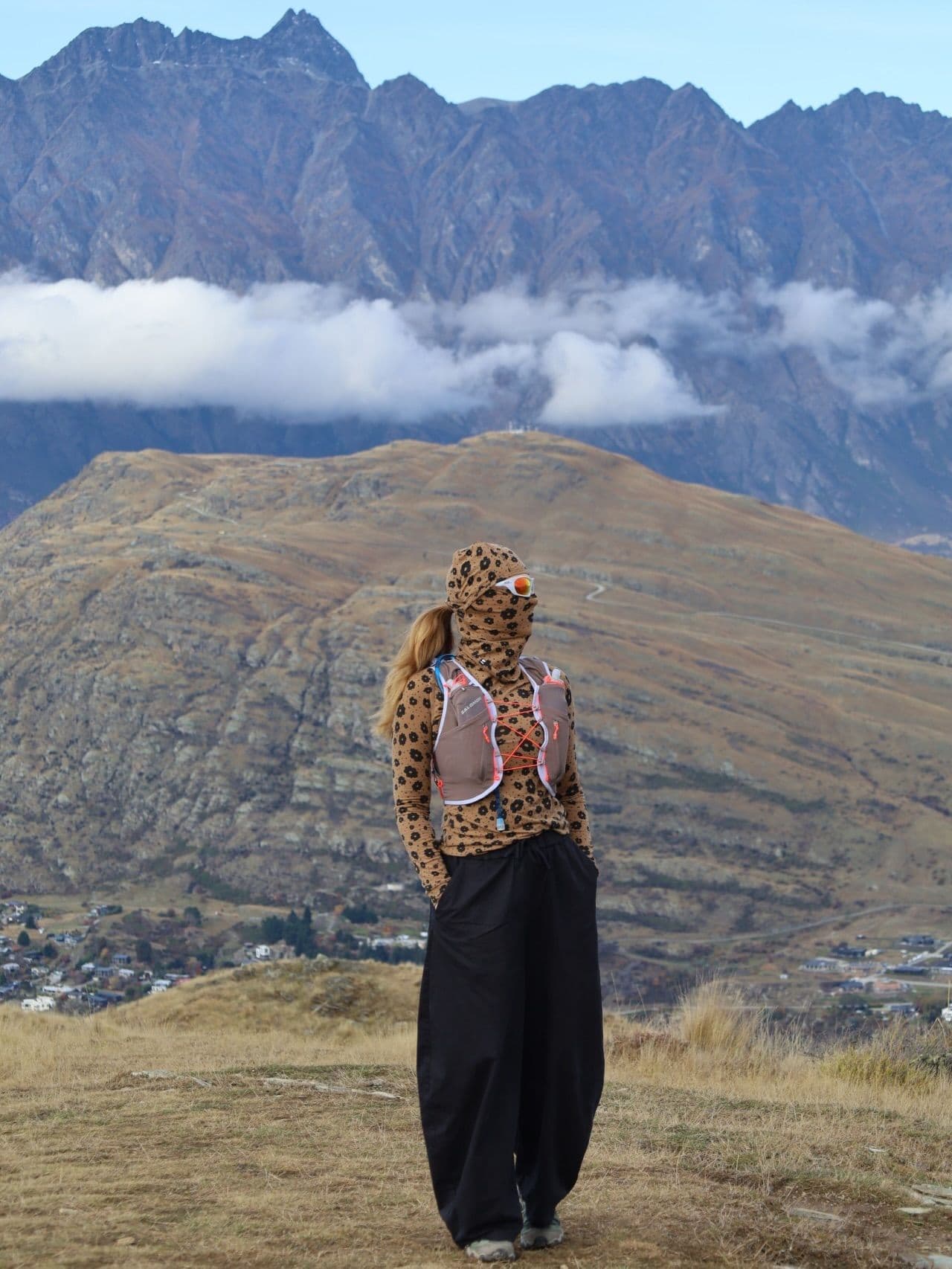 A woman in a leopard print balaclava, sunglasses, and running vest stands on a dry hill overlooking a valley town and distant mountains.