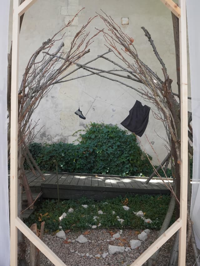 An outdoor art installation with a wooden frame surrounding an arch of bare branches, from which a single shoe and a black top hang above a garden path.