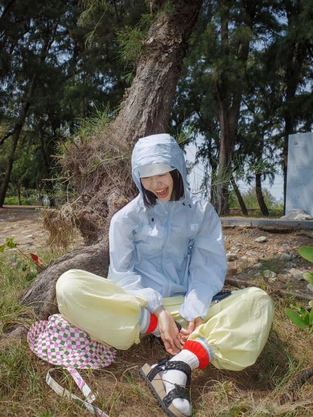 A person in a light blue hooded jacket and yellow pants sits smiling by a tree, with a checkered hat on the ground next to them.