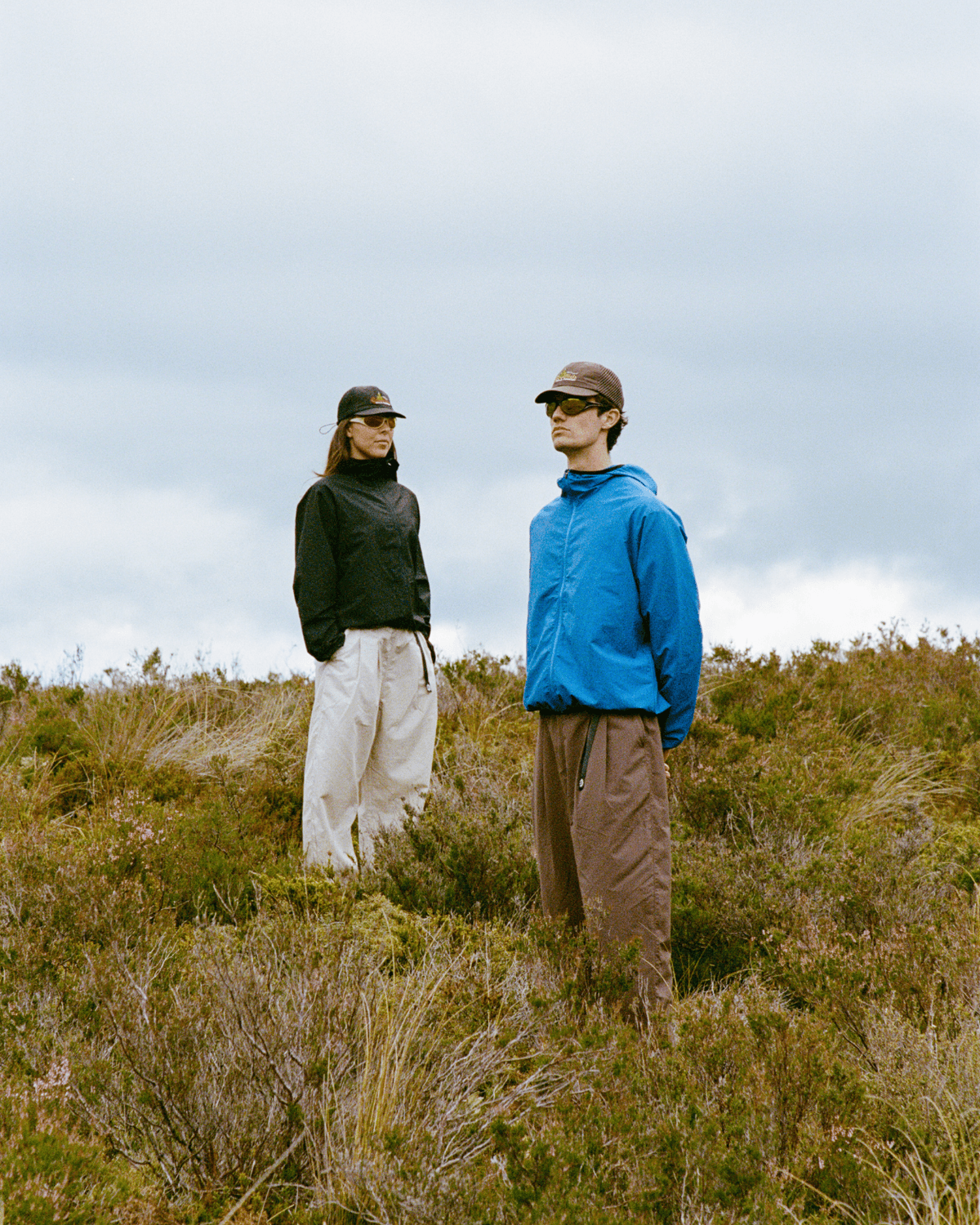 A man and a woman in outdoor gear stand in a field of dry grass and shrubs under a cloudy sky.