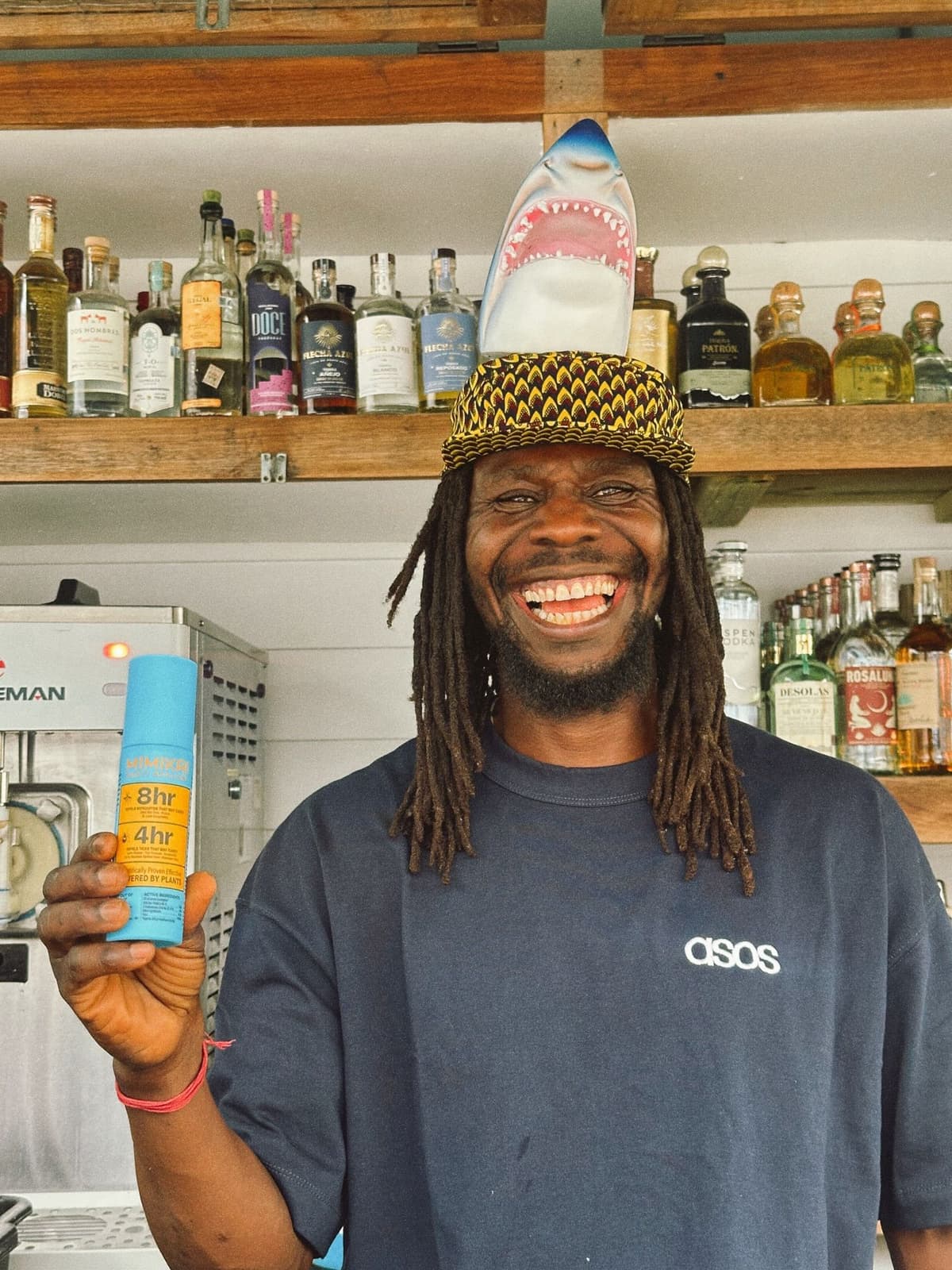 A smiling Black man with dreadlocks, wearing a shark-fin hat and an ASOS t-shirt, holds a blue spray bottle in front of a liquor bar.