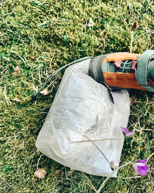 A translucent ultralight dry bag and a hiking sandal rest on vibrant green moss with small purple flowers.