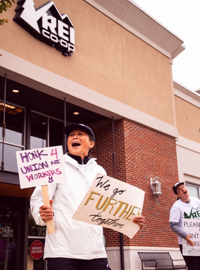 A woman shouts, holding "HONK 4 UNION REI WORKERS" and "We go FURTHER together" signs outside an REI Co-op store.