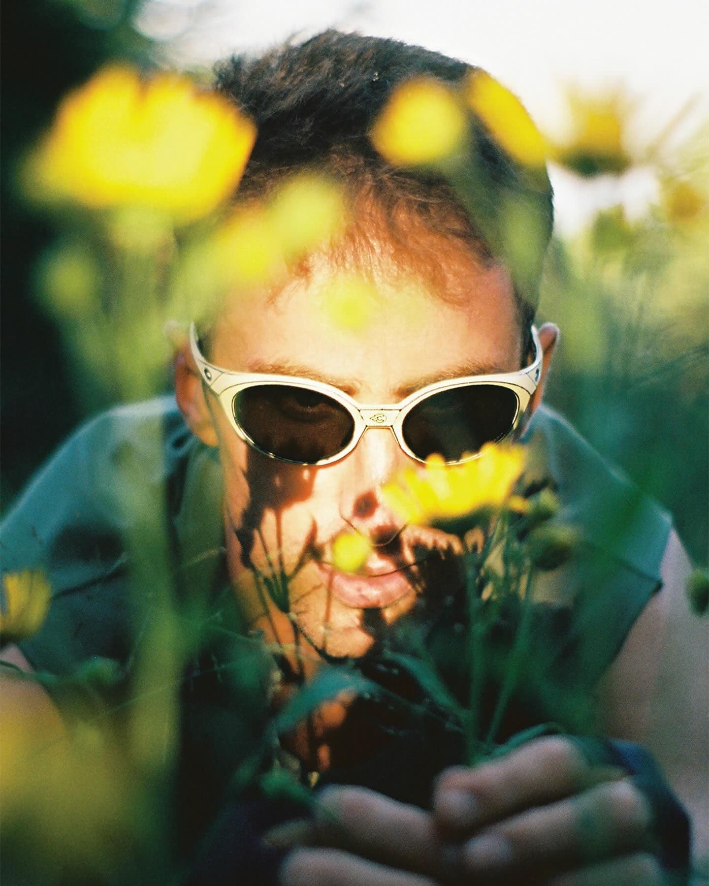 A person in white sunglasses peeking through blurred yellow flowers, holding green stems.