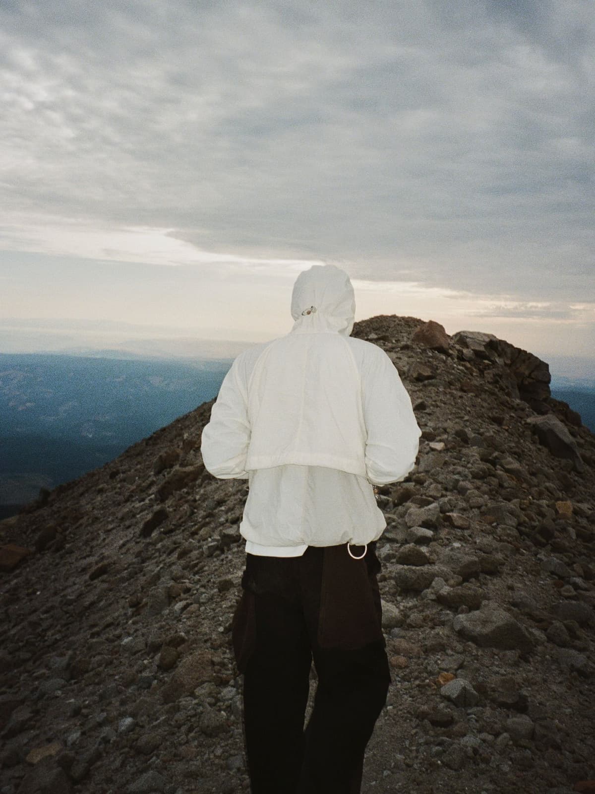 Person in a white hooded jacket walks on a rocky mountain ridge under a cloudy sky.